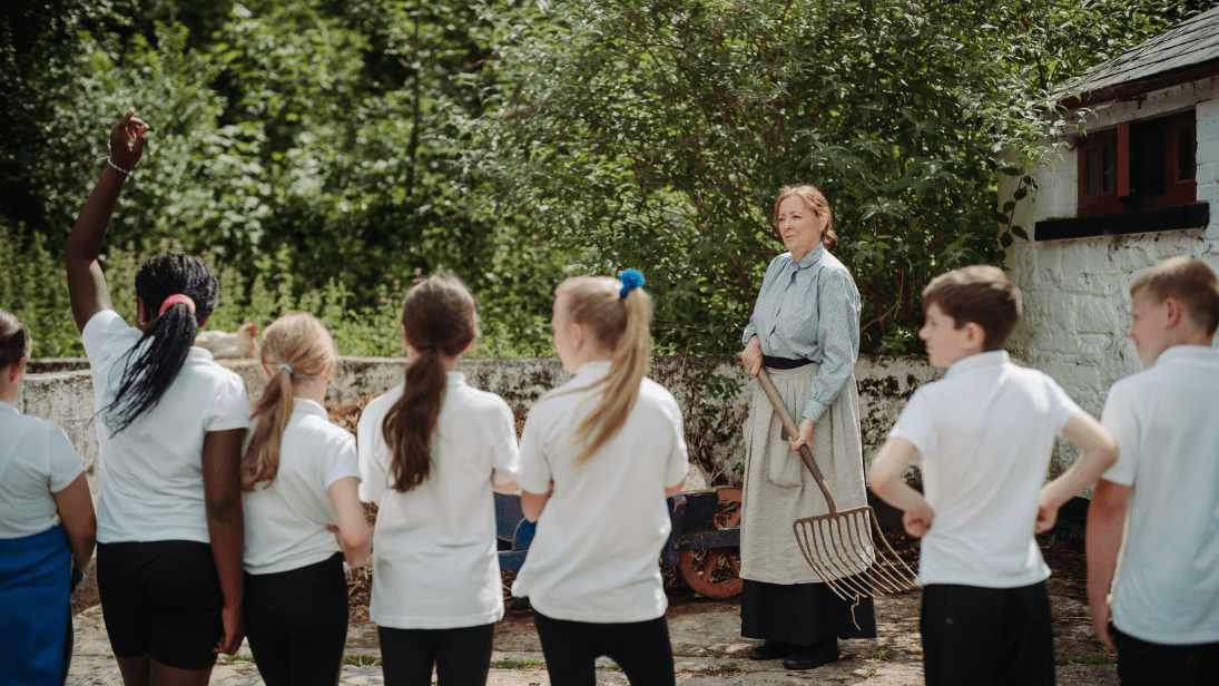 Costumed guide at the Ulster Folk Museum holding a pitch fork on the farm during a learning programme for schools. Seven school children dressed in white t-shirts all have their backs to the camera, as they're interacting with the guide.