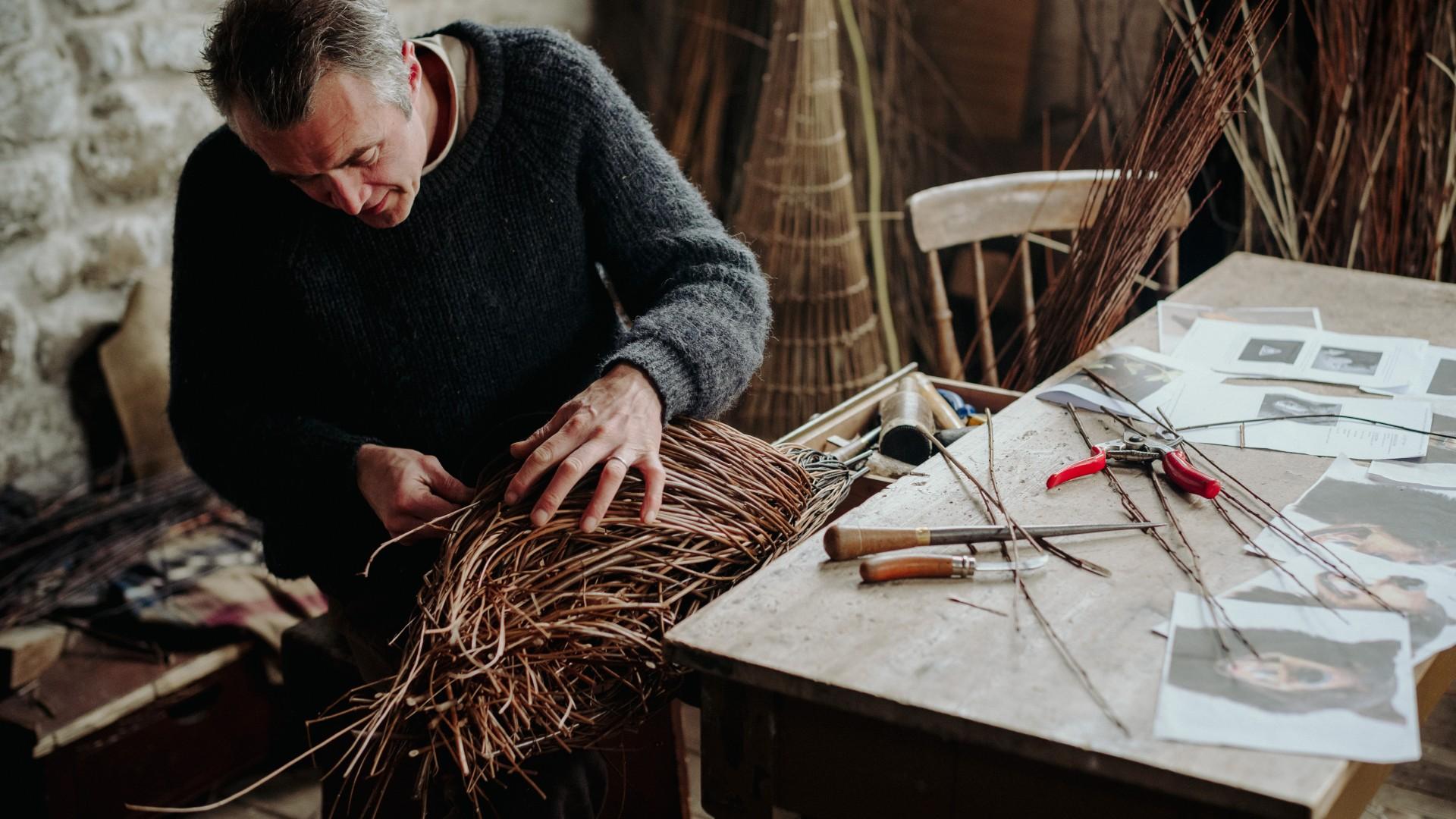 A man sits weaving willow together in a basket-like formation.