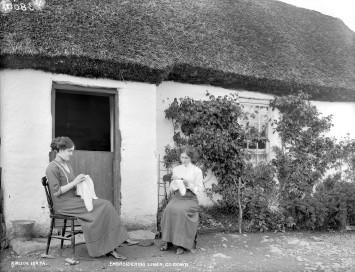 Two girls embroidering linen outside thatched house