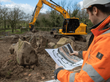 A man holds documents depicting the court tomb he is standing in front of. He wears a hard hat; in the background is a big digger.
