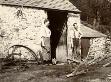 Two blacksmiths stand outside a forge, with many metal objects for agriculture around them.