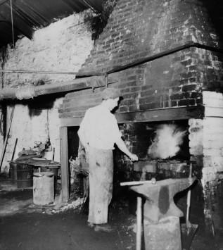 A man stands with his back to the camera. He is by the forge fire, holding a metal object.