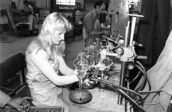 A woman sits at a work desk, working with blown glass.