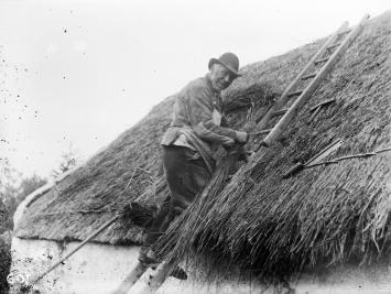 A man on a ladder turns towards the camera. He is thatching the roof of a cottage.