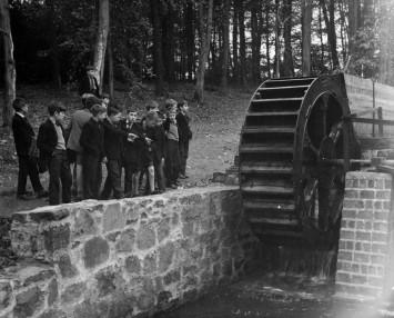 A black and white image of a school group looking at a water wheel.