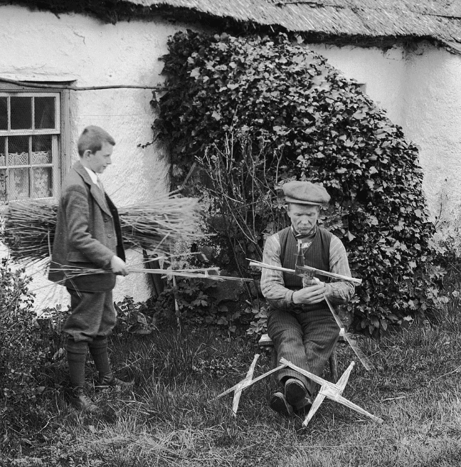 A black and white old photograph with two men outside of a cottage. One is standing holding rushes, the other sits and makes St Brigid's crosses.