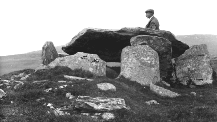 A man stands behind a dolmen, an archaeological monument of stones.
