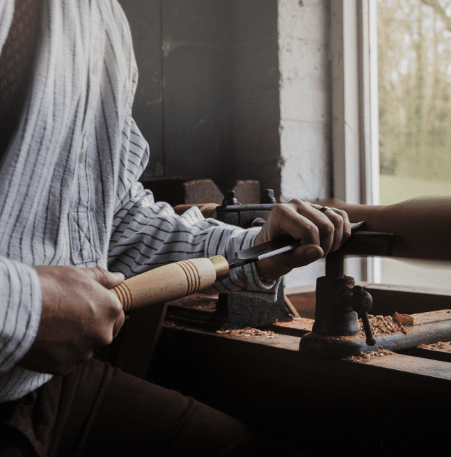 A carpenter uses a tool on a foot-turned lathe as a piece of wood spins.