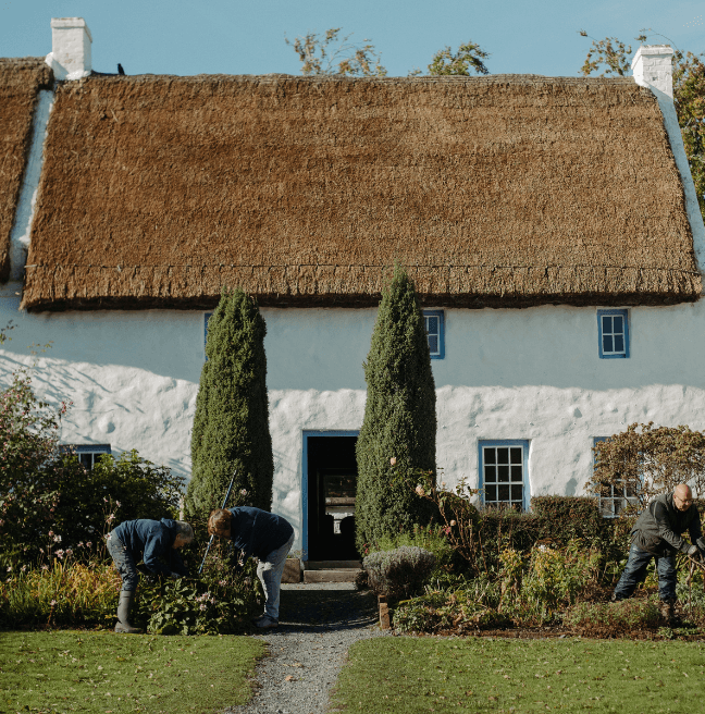 Three people work in the garden of a large, white thatched house.