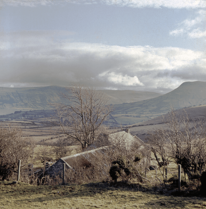 A colour photograph. In the foreground, a farmhouse sits on a hill. In the background, the Glens of Antrim stretch into the distance on a sunny day.