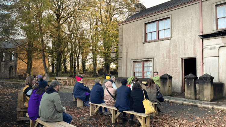 People sitting on benches outdoors at Ulster Folk Museum listening to storyteller