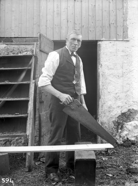A very tall man stands posing for the camera holding a large saw over a plank of wood.